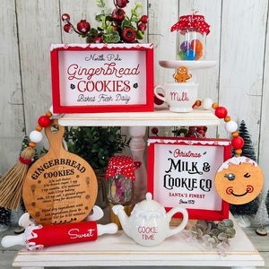 May include: A tiered white shelf display decorated for Christmas with red and white accents. The top shelf features a red sign that reads "North Pole Gingerbread Cookies Baked Fresh Daily", a glass jar filled with colorful candies, a white ceramic mug with the word "Milk", and a gingerbread man figurine. The middle shelf features a wooden cutting board with a gingerbread cookie recipe, a red rolling pin with the word "Sweet", a small white teapot with the words "Cookie Time", and a glass jar filled with colorful candies. The bottom shelf features a red sign that reads "Christmas Milk & Cookie Co. Santa's Finest Since 1872", a wooden gingerbread man ornament with a smiling face, and a small green potted plant.