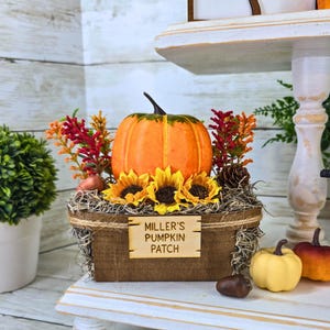 May include: A wooden box with a sign that reads "Miller's Pumpkin Patch" is filled with hay, a small orange pumpkin, and yellow sunflowers. The box is sitting on a white shelf with a small green bush and a white pot.