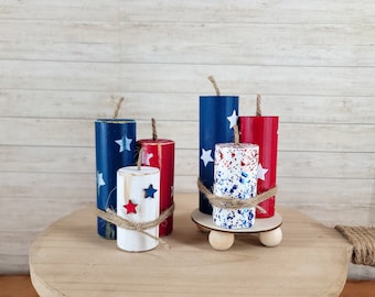May include: Wooden firecracker decorations in red, white, and blue with star accents. The patriotic decor includes a small wooden platform with wooden balls as feet, and is tied with twine. The background is a light wood.