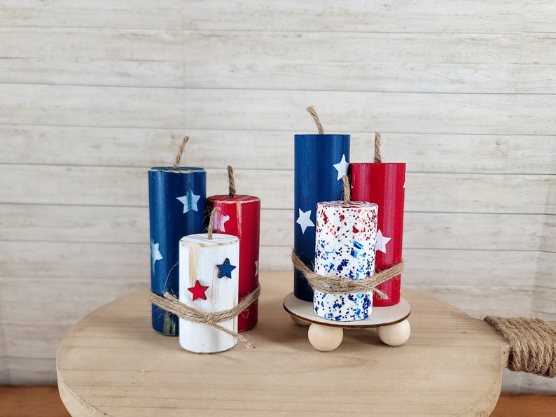 May include: Wooden firecracker decorations in red, white, and blue with star accents. The patriotic decor includes a small wooden platform with wooden balls as feet, and is tied with twine. The background is a light wood.