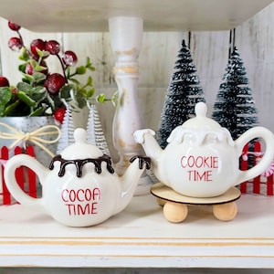 May include: Two white ceramic teapots with red lettering. One teapot says "Cookie Time" and the other says "Cocoa Time". The teapots are on a white tiered tray with a wooden base.