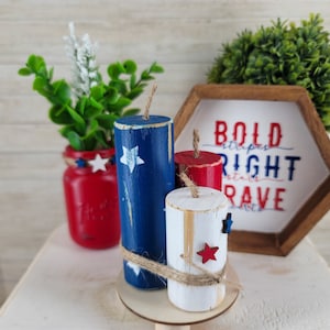 May include: Patriotic wooden firecracker decorations in red, white, and blue with star accents. The display includes a red mason jar with greenery and a wooden sign that reads "Bold Stripes, Right Stars, Brave."