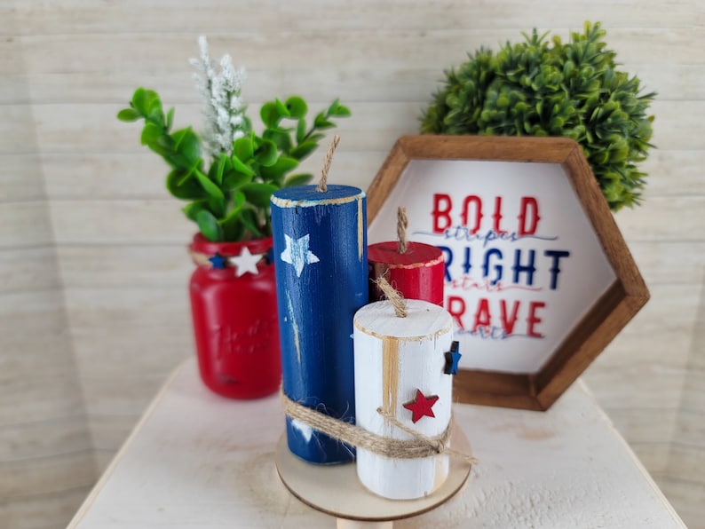 May include: Patriotic wooden firecracker decorations in red, white, and blue with star accents. The display includes a red mason jar with greenery and a wooden sign that reads "Bold Stripes, Right Stars, Brave."