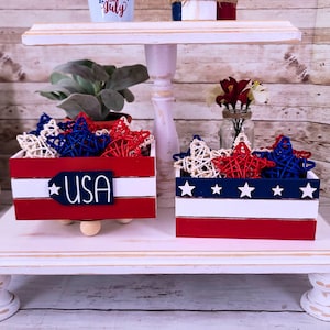 May include: A white tiered tray with two red, white, and blue wooden boxes filled with red, white, and blue star-shaped decorations. The boxes have the words "USA" and stars printed on them.