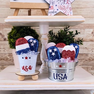 May include: Two small white buckets with red, white, and blue glitter popsicle decorations. The buckets are labeled "USA" and are sitting on a white tiered shelf.