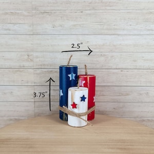 May include: Three wooden firecracker decorations painted in red, white, and blue with star accents. The tallest is 3.75 inches high. The firecrackers are tied together with twine and are 2.5 inches wide.