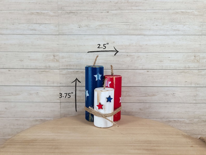 May include: Three wooden firecracker decorations painted in red, white, and blue with star accents. The tallest is 3.75 inches high. The firecrackers are tied together with twine and are 2.5 inches wide.