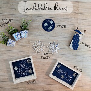 May include: A set of winter-themed decorations including a wooden sled, two framed signs with the text "Let it Snow" and "Feeling Frosty", three small potted plants, and two snowflake ornaments. The signs are painted blue with white lettering and snowflakes. The sled is painted blue and white. The plants are in white pots with blue speckles.