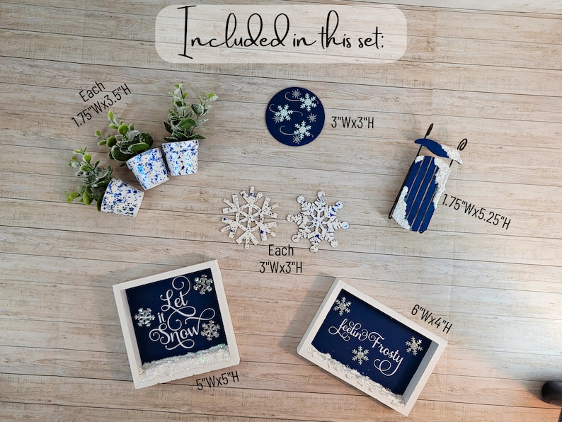 May include: A set of winter-themed decorations including a wooden sled, two framed signs with the text "Let it Snow" and "Feeling Frosty", three small potted plants, and two snowflake ornaments. The signs are painted blue with white lettering and snowflakes. The sled is painted blue and white. The plants are in white pots with blue speckles.