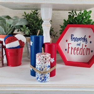 May include: A tiered shelf displays patriotic decor. Items include red, white, and blue candles, a red bucket with star-spangled ornaments, and a red and white hexagon sign that reads "Fireworks and Freedom."