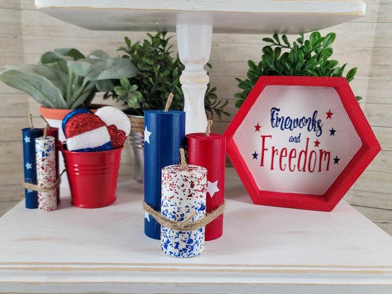 May include: A tiered shelf displays patriotic decor. Items include red, white, and blue candles, a red bucket with star-spangled ornaments, and a red and white hexagon sign that reads "Fireworks and Freedom."