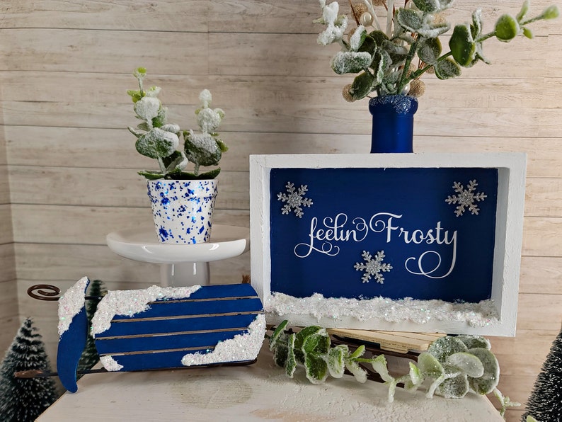 May include: A blue and white wooden sign with the text "Feelin' Frosty" in white cursive lettering. The sign is decorated with white snowflakes and a white snowy border. The sign is sitting on a white surface with a blue and white planter and a blue and white sled.