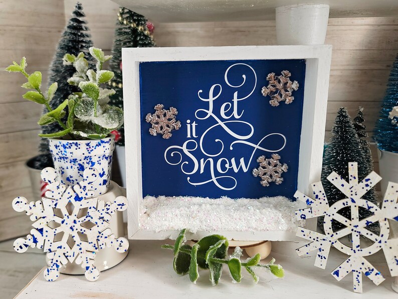 May include: A white wooden frame with a blue background and the text "Let it Snow" in white cursive lettering. There are silver glitter snowflakes on the blue background and a white, sparkly, snow-like substance at the bottom of the frame.