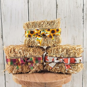 May include: Three miniature hay bales decorated with colorful ribbon. The top bale has a sunflower ribbon, the middle bale has a red, green, and yellow plaid ribbon, and the bottom bale has a white ribbon with orange pumpkins and leaves.