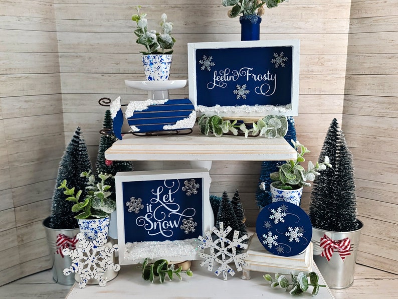 May include: A winter-themed tiered tray display with blue and white decorations. The top tier features a blue and white sled with a white sign that says "Feelin' Frosty". The middle tier has a blue and white sign that says "Let it Snow" and a white snowflake ornament. The bottom tier has a blue and white round ornament with snowflakes and a white snowflake ornament.
