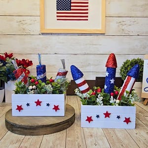 May include: Two white wooden boxes decorated with red and blue stars, each containing faux greenery and glittery firework decorations. An American flag is framed in the background, suggesting a patriotic theme.