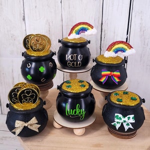 May include: A collection of black miniature pots filled with gold glitter and coins, and topped with rainbows. Some pots have the words "Pot O' Gold" or "lucky" on them, and are decorated with shamrocks and bows. The pots are displayed on small wooden stands.