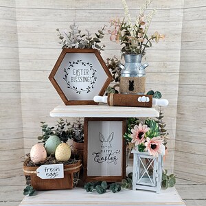 May include: A two-tiered white wooden shelf with Easter decorations. The top shelf has a hexagonal wooden sign with the text "Easter Blessings", a metal pitcher with a burlap bunny, and a wooden rolling pin. The bottom shelf has a wooden sign with the text "Happy Easter", a basket of pastel colored eggs, a white lantern, and a bouquet of pink flowers.