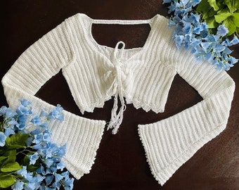 May include: White crochet crop top with long sleeves and a tie at the front. The top has a scalloped edge and a loose fit.