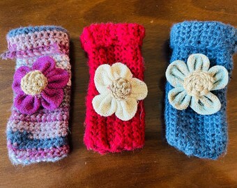 May include: Three handmade crochet cozies in shades of pink, red, and blue, each adorned with a fabric flower. The cozy on the left is striped with a purple flower, the middle one is red with a white flower, and the right one is blue with a light blue flower.