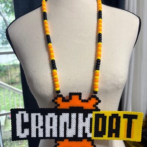 May include: A necklace featuring a rectangular pendant made of black, white, and yellow plastic beads arranged in a pixelated style with the text CRANKDAT. The pendant is attached to a long strand of alternating black, orange, and yellow pony beads.