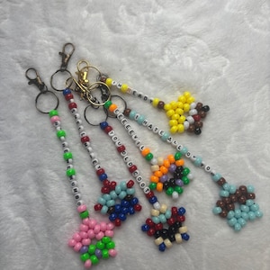 May include: A collection of beaded keychains in various colors. Each keychain features a unique design, including floral shapes and phrases like "I'M READY" and "I HATE ALL OF YOU". The keychains have metal clasps.