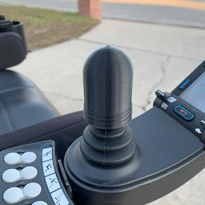 May include: A black joystick with a textured knob and a control panel with multiple buttons. The control panel has icons for seat adjustments.