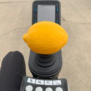 May include: A black joystick with a yellow lemon on top. The joystick has a screen and buttons with symbols for different functions.