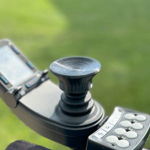 May include: A black joystick with a round, textured top and multiple buttons. The joystick is mounted on a black base.