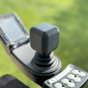 May include: A black joystick with a square-shaped top and multiple buttons. The joystick is mounted on a black base with a control panel.