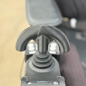 May include: Close-up of a black joystick controller with a curved handle. The controller is part of a mobility device, featuring a textured surface and a series of buttons and controls. The background is blurred, focusing attention on the device.