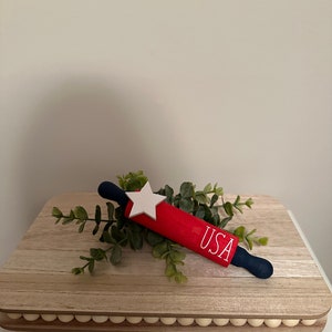 May include: Red and blue rolling pin with a white star and the word "USA" written on it. The rolling pin is on a wooden board with a beaded edge.