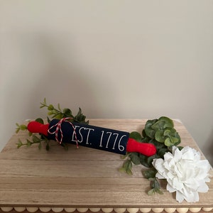 May include: A blue and red rolling pin with the text "EST. 1776" on a wooden surface with a white flower and green foliage.
