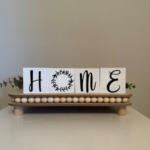 May include: White wooden blocks spelling "HOME" with a green wreath design on the "O" block. The blocks are sitting on a wooden stand with a beaded edge.