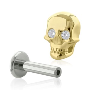 May include: A gold-toned skull-shaped stud earring featuring two clear, round, faceted stones set into the eye sockets. The piece includes a silver-toned flat-back post with a hollow cylindrical end, designed for secure piercing placement.