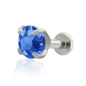 May include: A labret stud body jewelry piece featuring a round, faceted blue gemstone held in a four-prong silver-toned metal setting. The post is straight and terminates in a flat, circular disc backing, designed for secure placement in piercings.