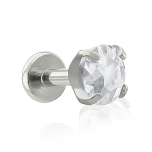 May include: A silver-toned labret stud featuring a round, clear, faceted gemstone held in a four-prong setting. The jewelry piece includes a flat circular back disc connected to a straight post, showcasing a polished metallic finish throughout.