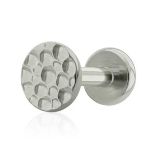 Puede incluir: Pendiente de botón plateado con textura martillada. El pendiente presenta una parte delantera plana y circular con un patrón irregular y con hoyuelos. El poste y la parte posterior del pendiente son lisos y cilíndricos.