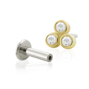 May include: A gold body jewelry piece with three clear round gemstones. The jewelry is designed to be worn in a piercing. The piece is shown next to a silver barbell.