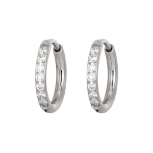 May include: A pair of silver-toned hoop earrings featuring a row of small, clear, round-cut stones embedded along the front-facing curve of each hoop. The earrings have a polished metallic finish and a hinged closure mechanism at the base.