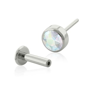 May include: A pair of surgical steel body jewelry with a clear iridescent gem. The gem is set in a round bezel. The jewelry is shown in two parts: the gem and the post. The post is a straight, smooth, cylindrical piece of metal with a small hole at one end.