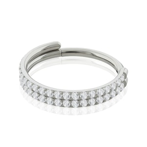 May include: A silver-colored hinged ring with two rows of clear, sparkling gemstones. The ring is shown against a white background, with a slight reflection visible below the ring.