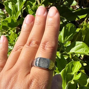 May include: A silver-toned signet ring featuring a smooth, flat, rectangular face and a polished metallic band. The ring has a solid construction with a reflective surface that catches the light, showcasing a clean, unadorned design on the finger.