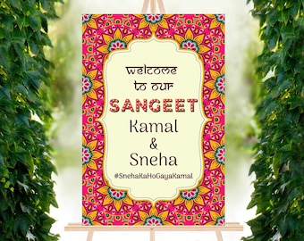 May include: A welcome sign for a Sangeet celebration with a pink, yellow, and blue mandala design. The sign reads "Welcome to our Sangeet Kamal & Sneha #SnehaKaHoGayaKamal".