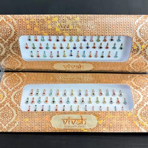 Puede incluir: Dos cajas de Vivah Designer Bindi, cada una con filas de bindis coloridos con tonos de joya. Los bindis están dispuestos en una bandeja con fondo blanco, sobre una caja dorada con un patrón ornamentado. El texto "Exclusive Wedding Bindi" es visible.