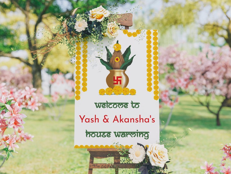 May include: A white sign with gold trim and yellow flowers welcomes guests to Yash & Akansha's housewarming. The sign features a traditional Indian pot with green leaves and a red swastika symbol.