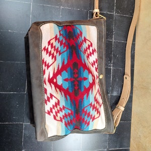 May include: A brown leather shoulder bag with a colorful geometric pattern on the front flap. The bag has a long adjustable strap and a small flap closure with a brass button. The bag is made by "SURI LEATHER COMPANY".