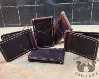May include: Six brown leather money clips with white stitching. The clips are arranged in a semi-circle on a wooden surface. The clips are all closed and have a metal clip on the front. The clips are all the same size and shape. The text "Stubby Leather Company" is visible in the bottom right corner of the image.