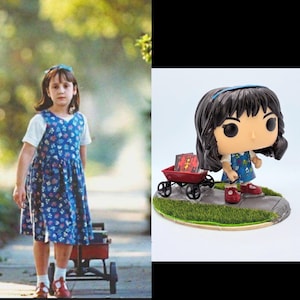 Può includere: Una statuetta Funko Pop di una ragazza con un vestito blu a fiori, scarpe rosse e una fascia blu, che tira un carretto rosso con libri. La statuetta è su una base con erba e un marciapiede.