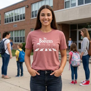 May include: A dusty rose short-sleeve t-shirt featuring a white graphic of four ghosts walking across a crosswalk with the text The Boosties printed above the illustration in a stylized white font, paired with dark blue denim jeans.